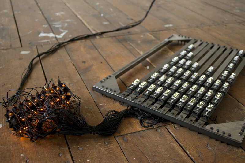 A detail of a metal frame laying on the wooden floor. Across it many small white numeric displays are brightly lit. Next to it a black structure with orange LEDs piercing through is connected to many small cables.
