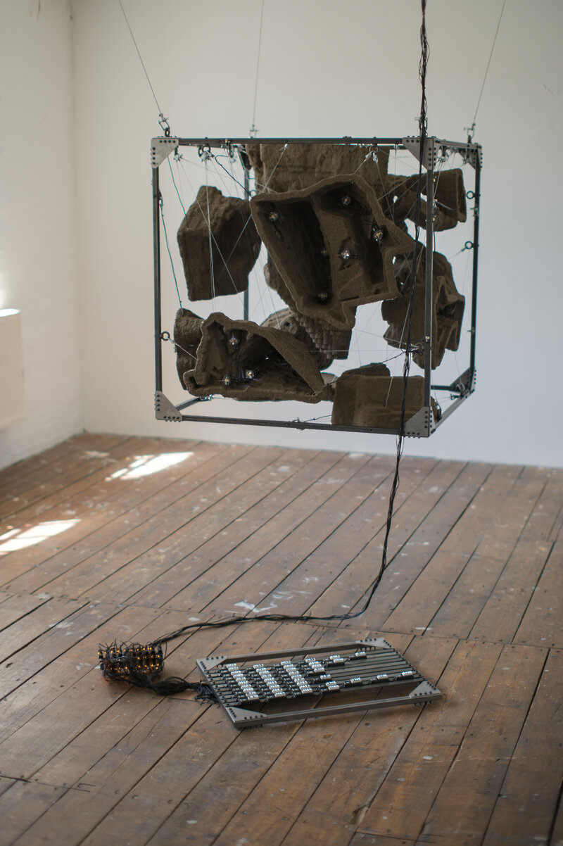 A metal frame in the shape of a cube is hanging from the ceiling. Inside of it structures made from earth resembling open-pit mines are suspended. In front of the cube a frame with small, white displays lays on the floor, a cable connecting both.