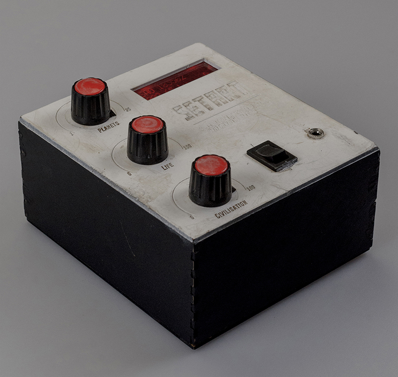 A black box with a white cover, a red LCD display, three rotating knobs and a 3.5mm audio jack. The cover looks worn and has scratches all over it.