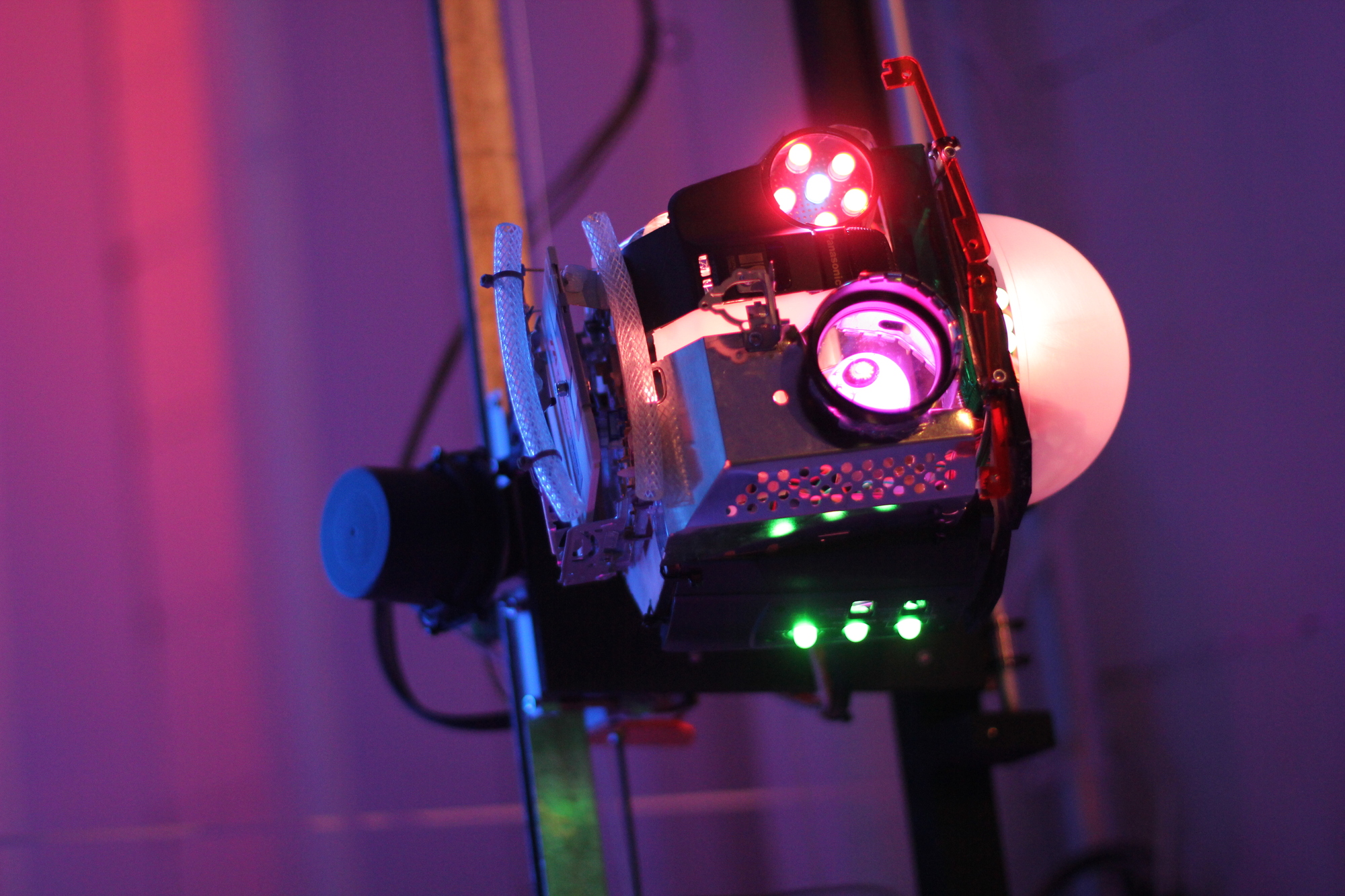 A robotic head made out of scrap, suspended from a rail in front of a pink-magenta illuminated background. Its left eye in from of an eyeball is shining magenta whilst its right eye is a ring of six red leds. Its eyebrows make it look surprised with its mouth slightly opened. Green lights pierce through the pierced-together structure.